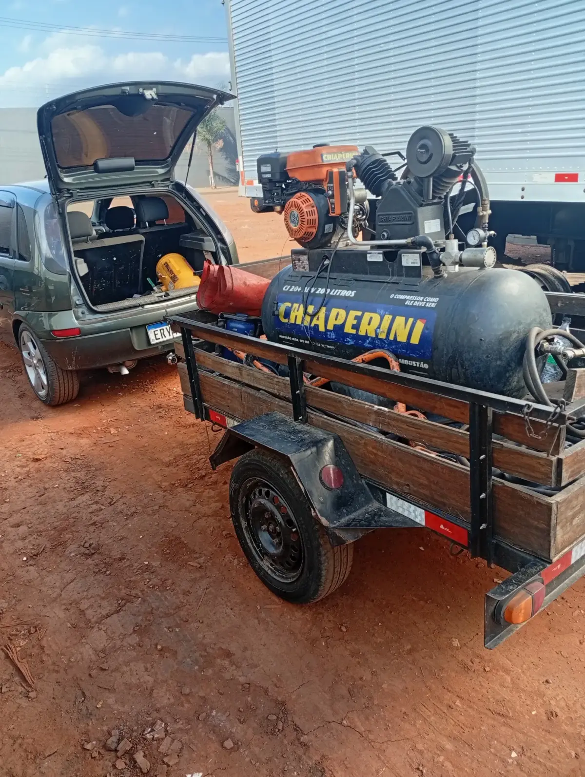 o carro de porta-malas aberto, engatado na carretinha de madeira com o compressor Chiaperini azul e o gerador laranja